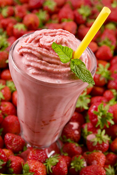 a strawberry milkshake in a tall milkshake glass garnished wtih a sprig of mint. against a background of strawberries