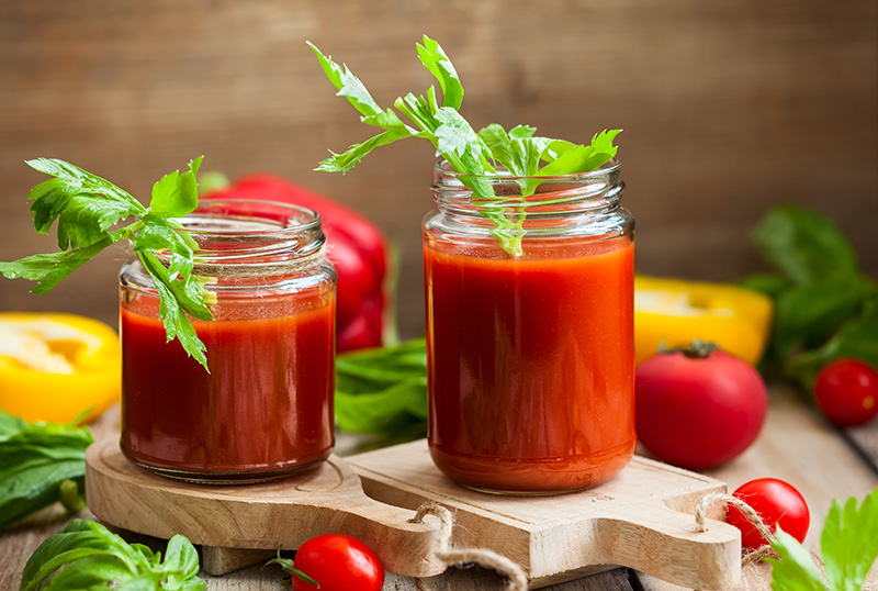 two glasses of tomato juice garnished with celery and surrounded by various vegetables