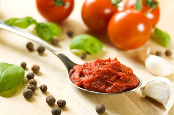 a spoon of tomato paste next to garlic cloves, peppercorns, basil leaves, and tomatoes