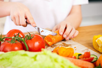 the hands of a child, seen cutting a carrot, with other vegetables nearby