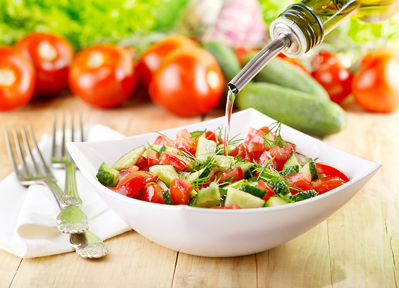 pouring olive oil onto a green salad of cucumbers and tomatoes