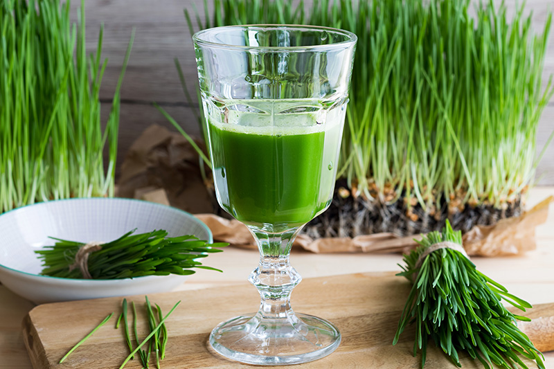 a glass of wheatgrass juice along with raw wheatgrass on a wooden table
